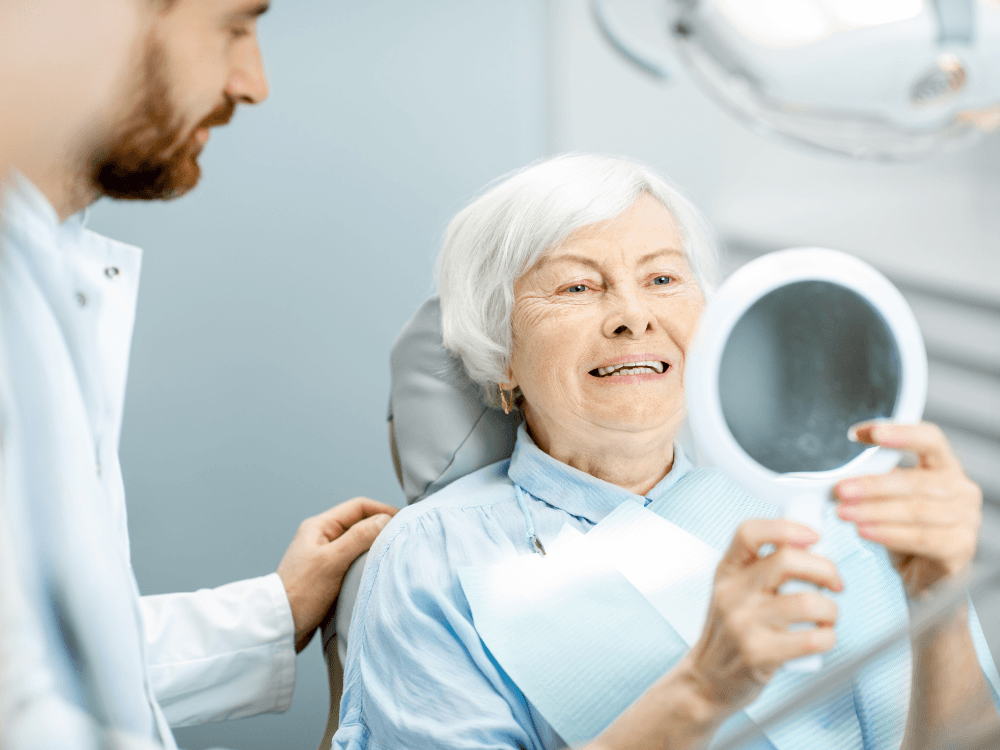Idosa observa o sorriso no espelho após avaliação para implante dentário sem parafusos acompanhada por cirurgião-dentista.
