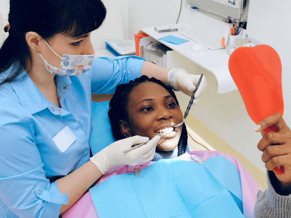 Dentista realiza exame clínico na boca de uma mulher negra deitada na cadeira odontológica, utilizando espelho e instrumentos dentários.