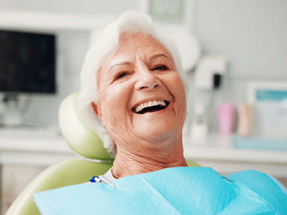 Idosa sorrindo sentada em cadeira odontológica, usando avental azul durante atendimento para colocação de implante dentário. Tudo para explicar quanto tempo demora para fazer um implante dentário.