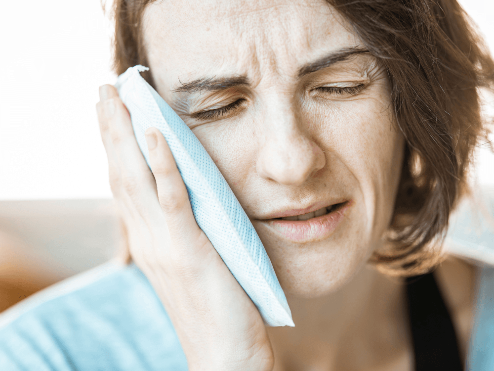 Mulher com expressão de desconforto aplica uma compressa fria na bochecha, representando o cuidado com a dor após implante dentário.