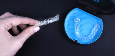 Woman holding invisible transparent aligners invisalign or plastic orthodontic braces. Black package with a box, clear-plastic retainers on blue background