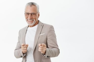 Het gotcha. Portrait of amused and happy carefree old man feeling energized ready to make money pointing at camera with finger gun gestures and smiling positive being in good mood over gray wall. Copy space